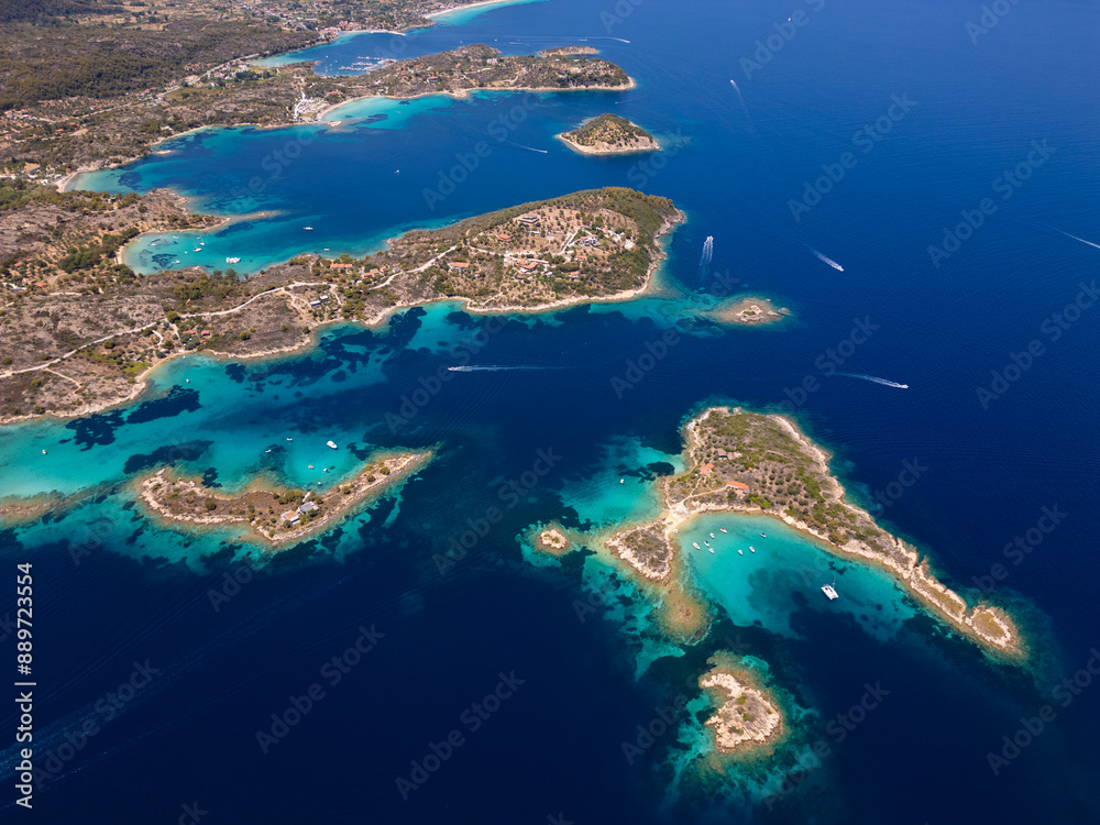Obraz premium Aerial view of an island on Sithonia Peninsula, Greece, with beaches and rocks, surrounded by crystal-clear waters. Yachts and boats anchor in turquoise waters. Nearby islets and reefs visible