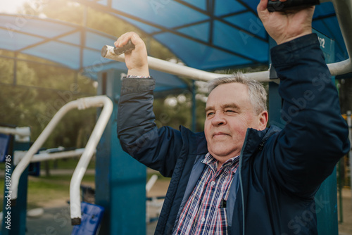 Senior man exercising outdoor fitness equipment in park. Elderly gentleman using pull-up bars under canopy. Active lifestyle and healthy aging concept.  photo