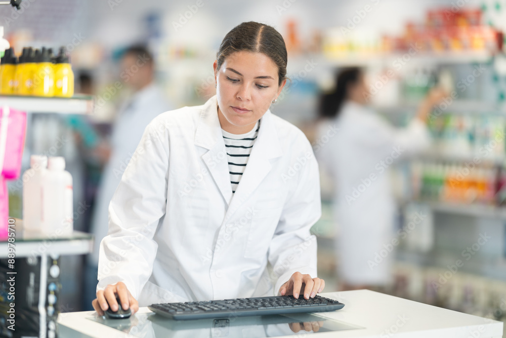 Fototapeta premium Young female pharmacist worker in uniform posing at computer in pharmacy