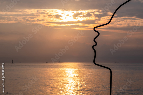 Cheongsapo, Haeundae-gu, Busan, South Korea - August 13, 2023: Silhouette and sunrise view of metal wire sculpture with human face shape near the sea at Daritdol Observatory
 photo