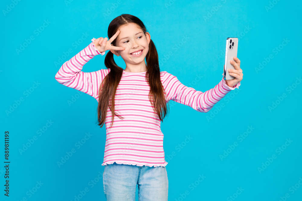 Photo of cute small girl with ponytails dressed striped shirt make selfie on smartphone show v-sign isolated on blue color background