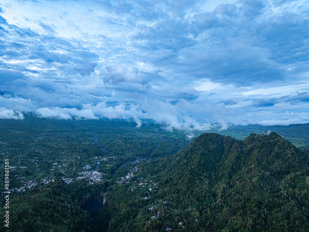 Aerial view Kabut Pelangi Waterfall in East Java. The waterfall stream flows into the mountain gorge in a long path.Above is the residence of the villagers. Abundance of forests on the green mountain. Aerial view Kabut Pelangi Waterfall in East Java. The waterfall stream flows into the mountain gorge in a long path.Above is the residence of the villagers. Abundance of forests on the green mountain.