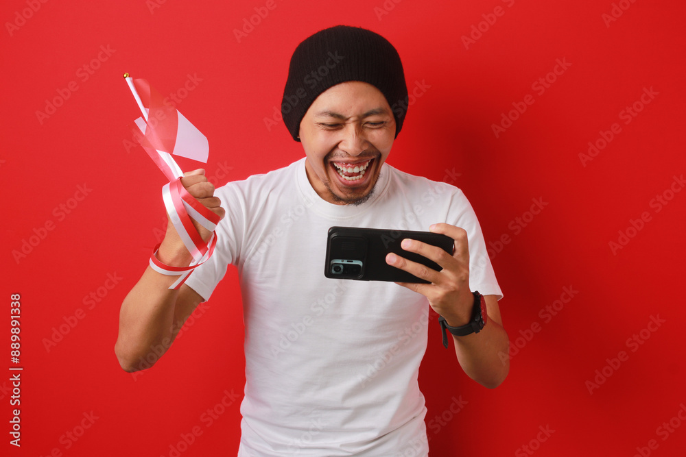 Overjoyed Indonesian man celebrates watching the Indonesia National team score a goal and win the game on his phone while holding the Indonesian flag during Indonesia Independence Day on August 17 Overjoyed Indonesian man celebrates watching the Indonesia National team score a goal and win the game on his phone while holding the Indonesian flag during Indonesia Independence Day on August 17