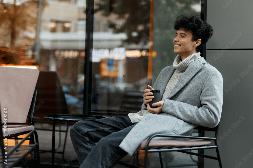 Young man sitting on a chair by a window, holding a cup of coffee with a smile on his face