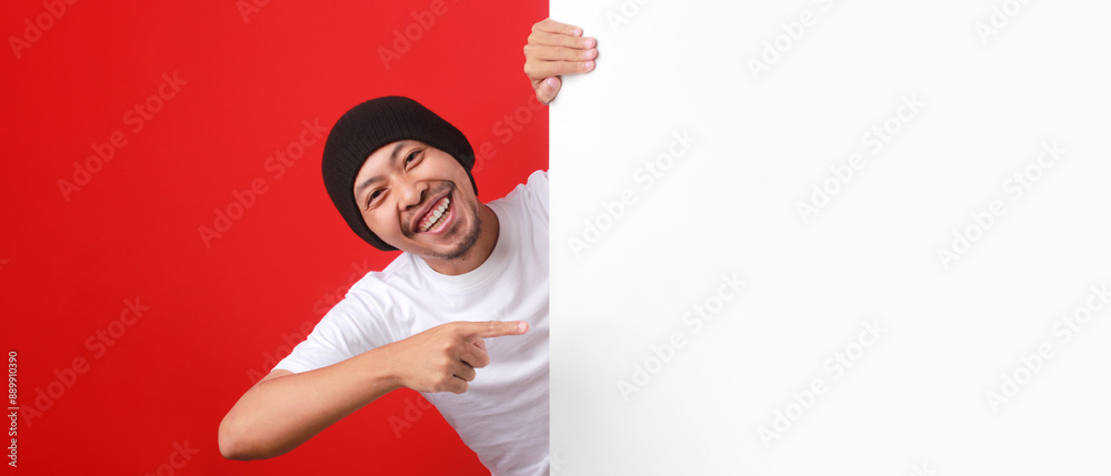 Indonesian man in a white T-shirt and beanie points at copy space, promoting a special offer or discount during Indonesia Independence Day. He stands behind a white wall, isolated on a red background. Indonesian man in a white T-shirt and beanie points at copy space, promoting a special offer or discount during Indonesia Independence Day. He stands behind a white wall, isolated on a red background.