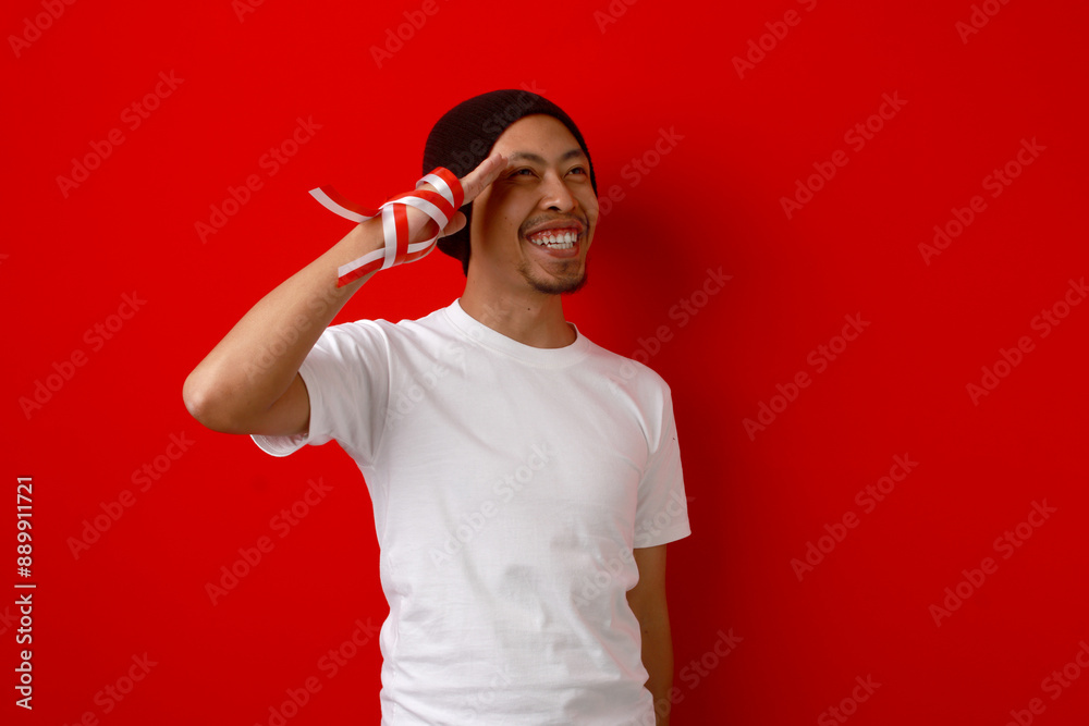 Fototapeta premium Young Indonesian man gives a salute gesture while celebrating Indonesian Independence Day on August 17, Isolated on a red background.