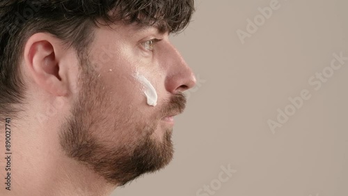 Young adult man applies aftershave cream moisturizer looks at camera, isolated on white background. Natural beauty skin care concept. Side view.