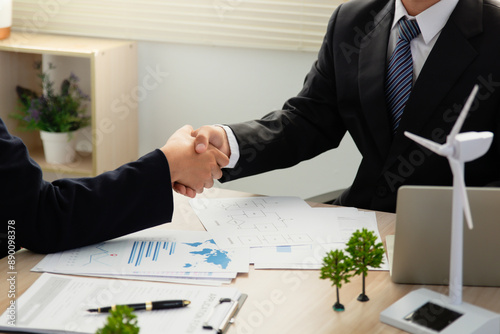 Green Business concept. Two people in suits shake hands over paperwork, a miniature wind turbine, and a small forest on a table. a deal or agreement related to renewable energy. ESG. Green energy. photo