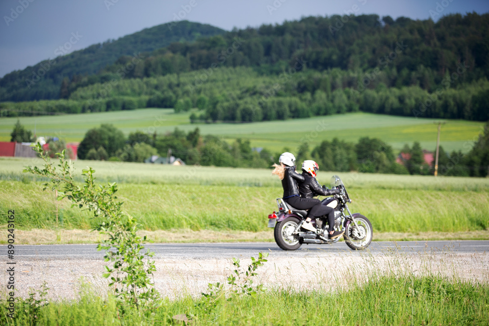 Motocykl na drodze z pasażerką, zielone pola, dachy domu, lasy na wzgórzu. Motocykl na drodze z pasażerką, zielone pola, dachy domu, lasy na wzgórzu.