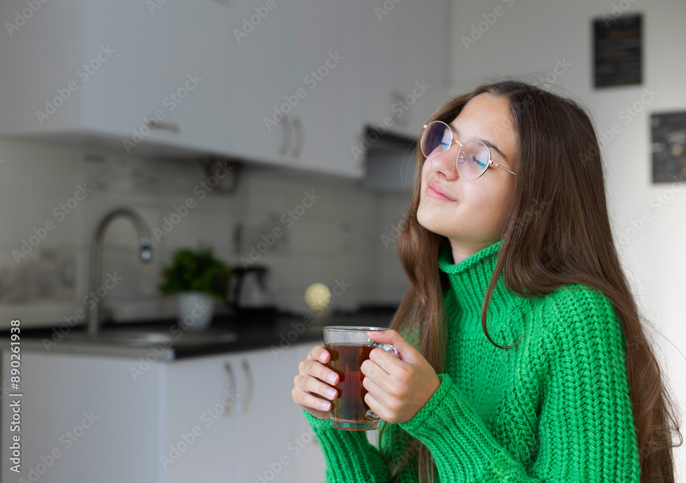 Relaxed young girl with glasses holding tea in the kitchen. Young woman with long brown hair wearing glasses and a green knitted sweater holding a clear glass cup of tea while smiling and closing her Relaxed young girl with glasses holding tea in the kitchen. Young woman with long brown hair wearing glasses and a green knitted sweater holding a clear glass cup of tea while smiling and closing her