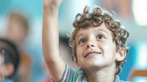 Curly-haired boy raising his hand in a classroom with a smile on his face. The background shows other children, indicating an engaging and interactive learning environment photo