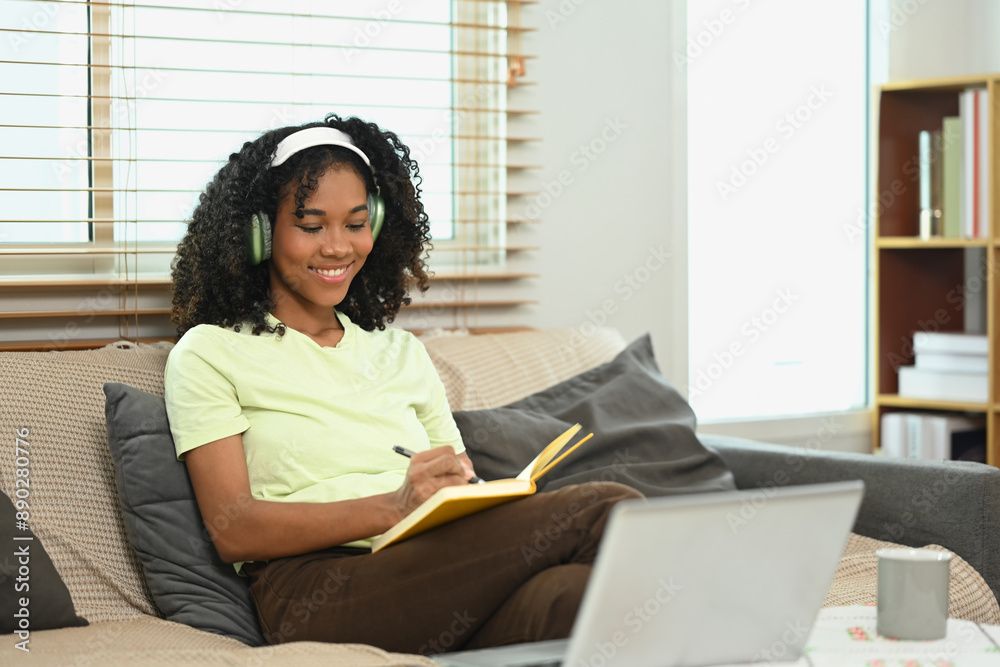 Pleased young Afro woman wearing headphone writing her diary on couch Pleased young Afro woman wearing headphone writing her diary on couch