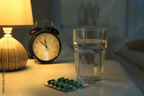 Insomnia treatment. Glass of water, pills and alarm clock on bedside table in bedroom at night photo