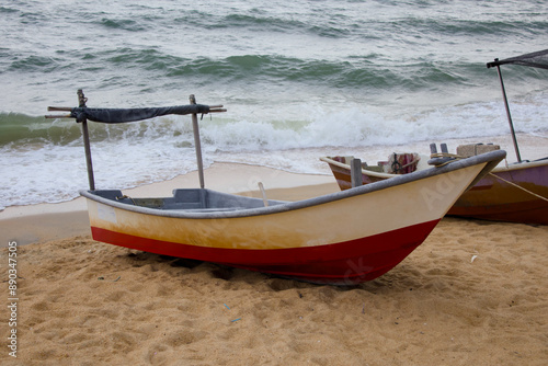 Fototapeta Naklejka Na Ścianę i Meble -  A vibrant red and white traditional fishing boat rests on the sandy beach of Pantai Teluk Lipat, Dungun, Terengganu. Equipped with wooden oars, it appears ready for a day at sea.