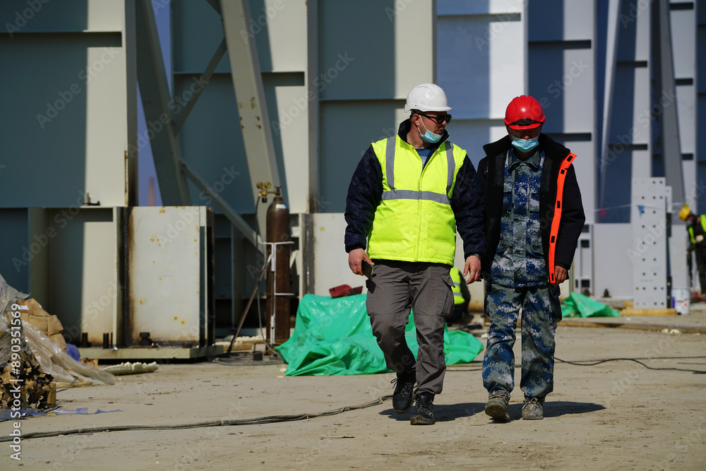 Two multiracial construction workers or engineers walking side by side through a construction site, conversing. Two multiracial construction workers or engineers walking side by side through a construction site, conversing.