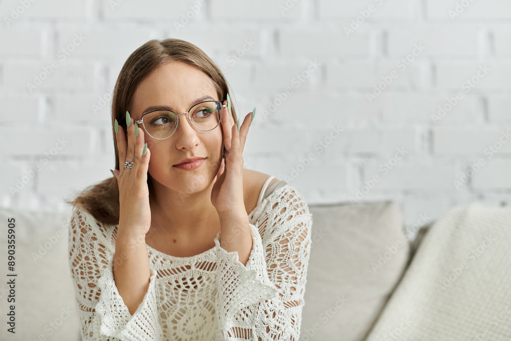 A young woman in a white lace top adjusts her glasses while sitting on a couch. A young woman in a white lace top adjusts her glasses while sitting on a couch.