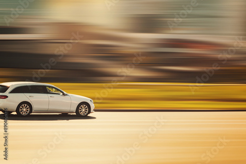 A white car is driving on an asphalt street on a sunny day with a blurred background at speed