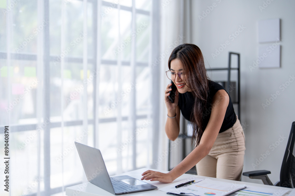 A woman is talking on her cell phone while sitting at a desk with a laptop A woman is talking on her cell phone while sitting at a desk with a laptop