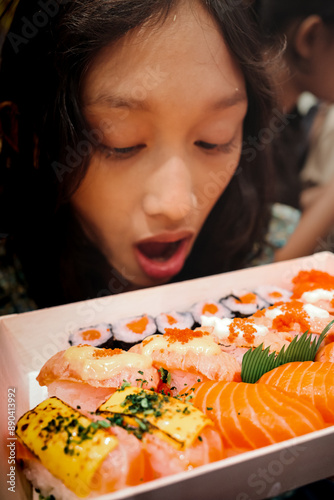 Southeast Asian teenage girl looking at a box of sushi and sashimi Japanese food bento meal desirously with her mouth wide opened photo