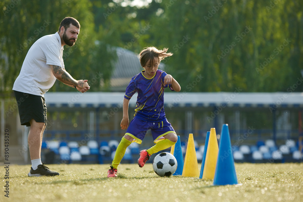 Fototapeta premium Young soccer player navigating ball through cones, while coach providing instruction and support. Outdoor football game. Concept of sport, childhood, education, achievement, active lifestyle