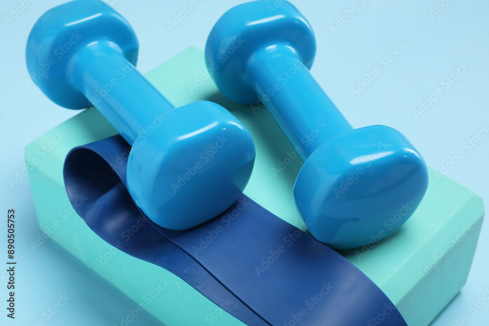 Two dumbbells, yoga block and fitness elastic band on light blue background, closeup