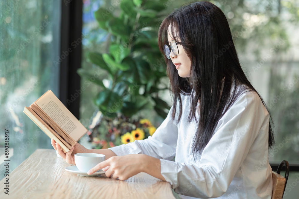 Obraz premium Young asian woman sitting in front of window relaxing in her living room reading book and drinking coffee or tea at home.