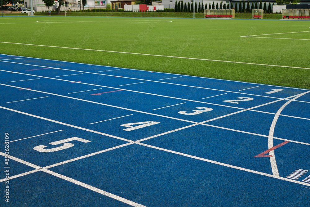 Fototapeta premium Treadmill with blue color and white numbers on it in the vohle of a large soccer field with green artificial grass. There are city buildings in the distance with nice trees growing between the houses.