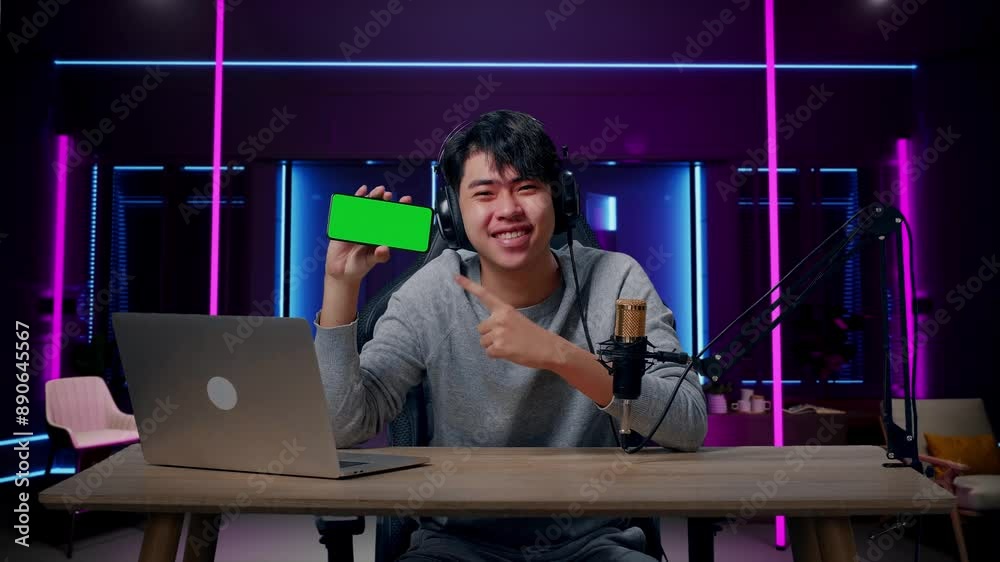A Smiling Asian Man With Headphone And Computer Showing Green Screen Mobile Phone In Room Lit By Neon Lights In Retro A Smiling Asian Man With Headphone And Computer Showing Green Screen Mobile Phone In Room Lit By Neon Lights In Retro