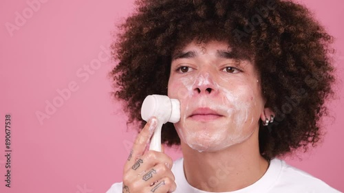 Young man using an electric facial brush for cleansing against a pink background.