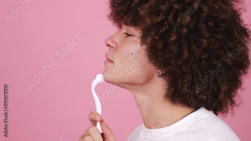 Close-up of a young man using a skincare derma roller on his face against a pink background.