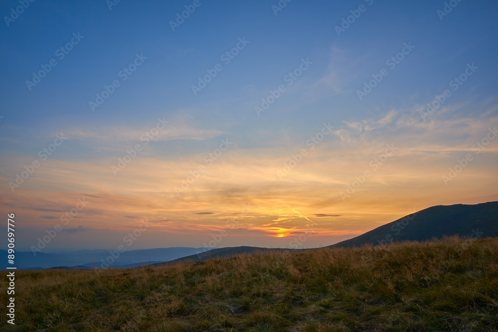 Obraz premium Sunset over grassy hills with a vibrant sky painted in shades of orange and blue, offering a tranquil and picturesque view of the distant mountains.