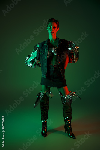 Woman in denim jacket and knee-high boots stands in studio, lit by green and red neon lights