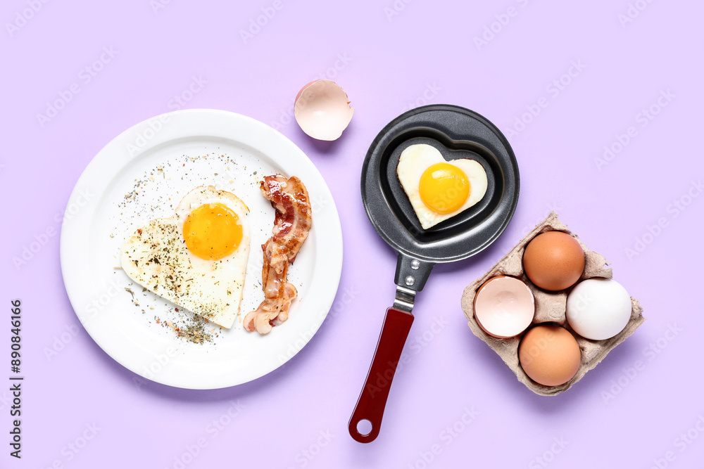 Composition with heart-shaped fried egg in frying pan and plate with fried egg and bacon on lilac background Composition with heart-shaped fried egg in frying pan and plate with fried egg and bacon on lilac background