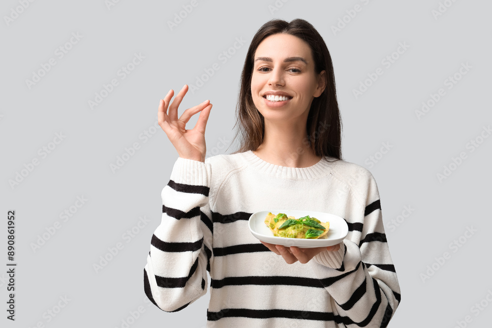 Beautiful young woman with plate of ravioli showing OK gesture on grey background Beautiful young woman with plate of ravioli showing OK gesture on grey background