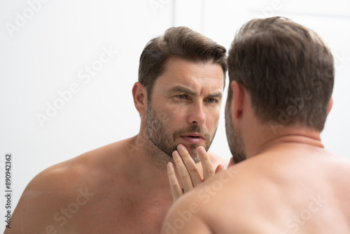 a man in a bathrobe in the bathroom looking at his wrinkles and pimples on his face. A man is looking at his reflection in a mirror. A man is looking at his reflection in a mirror. 