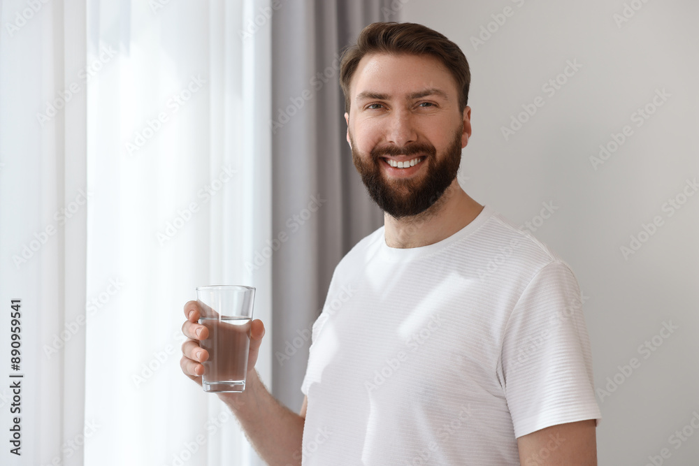 Happy young man with glass of water near window at morning