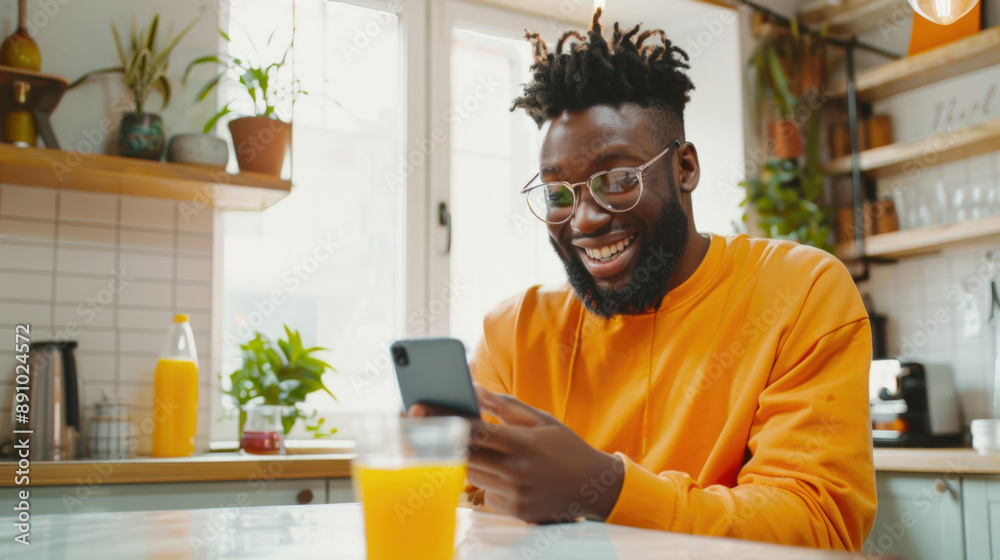 A black man in an orange shirt is sitting at a table with a glass of orange juice and a smartphone in his hands. He smiles and looks at his mobile phone, reads the morning news A black man in an orange shirt is sitting at a table with a glass of orange juice and a smartphone in his hands. He smiles and looks at his mobile phone, reads the morning news
