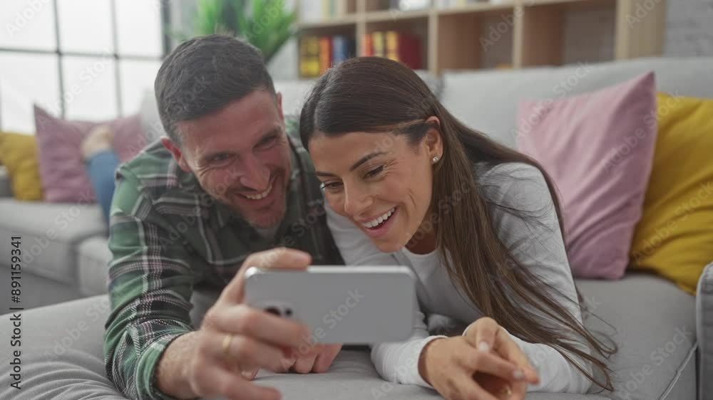 A smiling man and woman lying on a sofa interacting with a smartphone in a cozy living room. A smiling man and woman lying on a sofa interacting with a smartphone in a cozy living room.