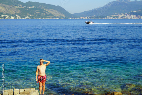 Beach holiday on the Adritic: a young man standing to his full height against the backdrop of mountains and the colorful sea. Tourist on the shore in the municipality of Herceg Novi, Montenegro. photo