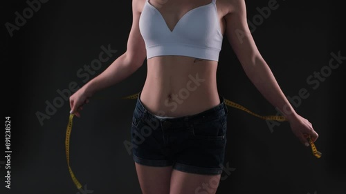 A slender girl in a beige white top and short black denim shorts measures her waist and hips with a measuring tape on a dark background. The concept of weight loss and healthy lifestyle.