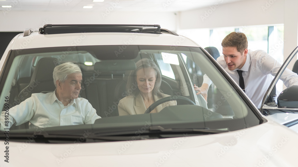 A gray-haired Caucasian woman is sitting behind the wheel of a car. Car dealership salesman consulting mature married couple.
