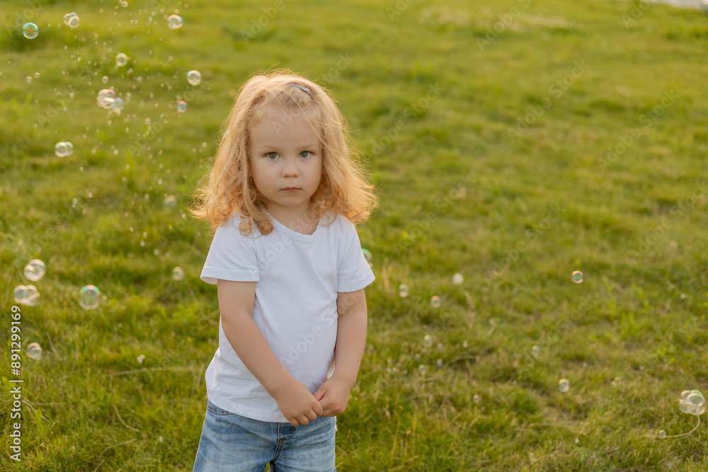 Cute blonde kid with curly hair walks on the lawn with soap bubbles in summer Cute blonde kid with curly hair walks on the lawn with soap bubbles in summer