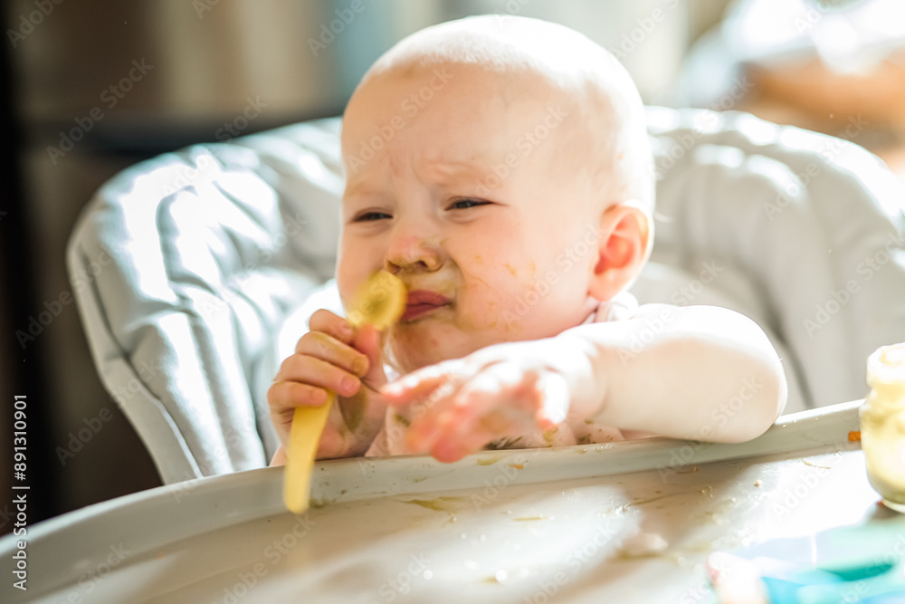 6 month Old Baby Girl At Home In High Chair eating tasty food at home. Funny baby with soiled healthy broccoli puree face holds spoon in hands 6 month Old Baby Girl At Home In High Chair eating tasty food at home. Funny baby with soiled healthy broccoli puree face holds spoon in hands