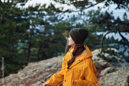 A woman in a vibrant yellow raincoat standing majestically on top of a mountain surrounded by lush green trees