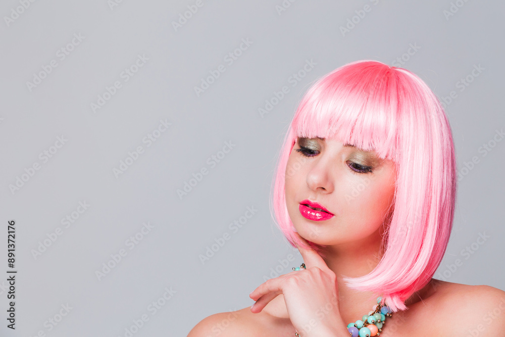 Young Caucasian woman with short pink hair, multi-colored necklace, and bare shoulders posing against a gray background. The woman has a serious expression and is looking down. Horizontal photo with c Young Caucasian woman with short pink hair, multi-colored necklace, and bare shoulders posing against a gray background. The woman has a serious expression and is looking down. Horizontal photo with c