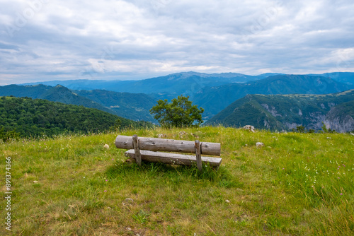 A log wooden bench stands on a hillside, with a green lawn around and mountains in the distance against a blue sky