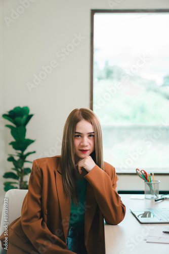 A woman in a brown jacket sits at a desk with a potted plant in the background. She is wearing a green shirt and has a red lipstick on. The scene suggests a professional setting