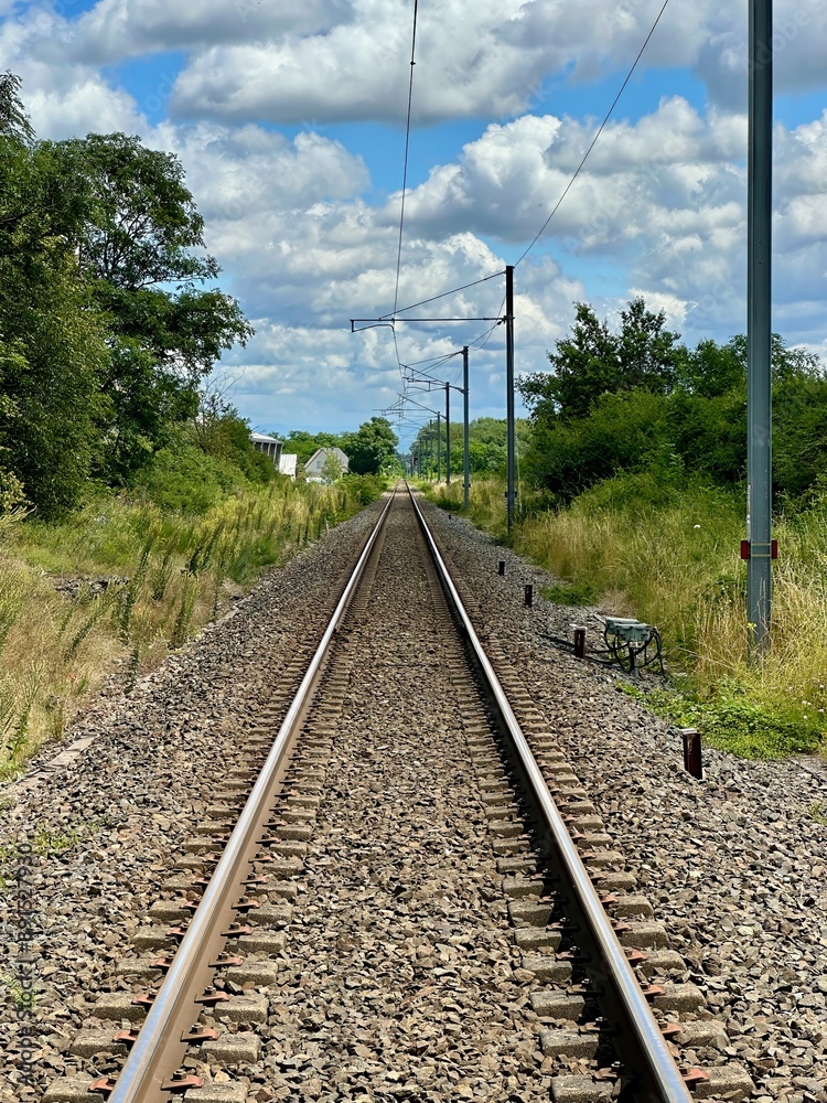 Long, Straight Railway Track Extending into the Distance Amidst Lush Greenery and Blue Sky with Fluffy Clouds Long, Straight Railway Track Extending into the Distance Amidst Lush Greenery and Blue Sky with Fluffy Clouds