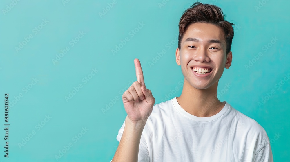 A cheerful young man in a white t-shirt points upwards, smiling brightly against a teal background, indicating enthusiasm and positivity.. A cheerful young man in a white t-shirt points upwards, smiling brightly against a teal background, indicating enthusiasm and positivity..