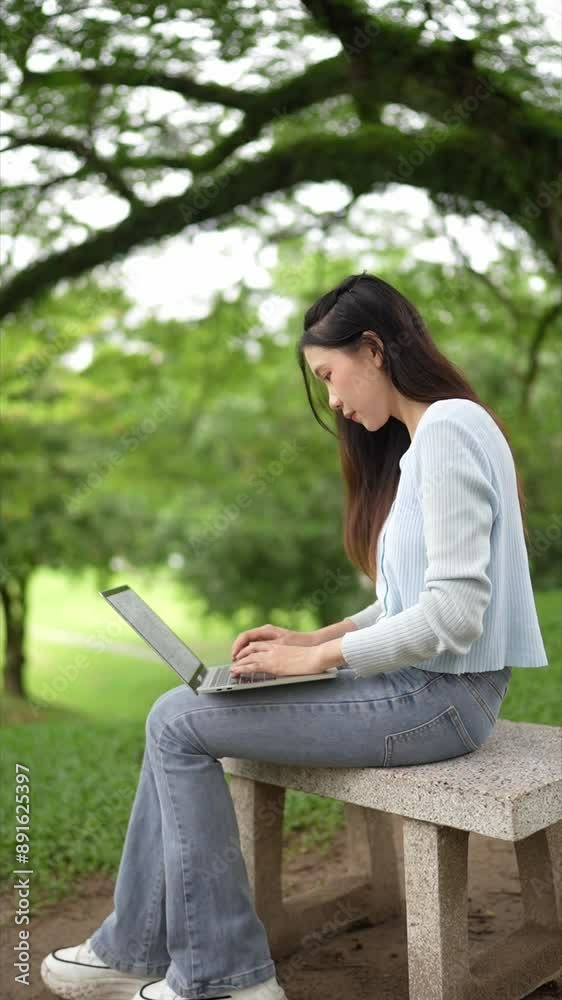 Asian woman sitting on a bench outdoors, working on a laptop. She is wearing a light blue top and jeans, surrounded by a green, lush environment Asian woman sitting on a bench outdoors, working on a laptop. She is wearing a light blue top and jeans, surrounded by a green, lush environment